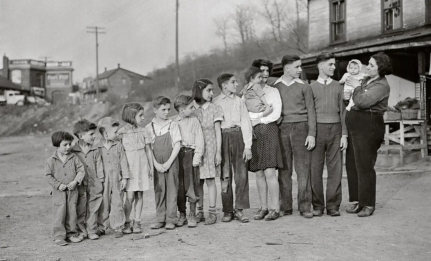 A family’s 13 children stand in increasing height, from small toddler girls and boys, to the tallest teenage sons. At the end stands the middle-aged Mrs. Celia Wega, who is also carrying a 3-month-old.